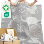 Glückliches Baby liegt unter grau-weißer gestrickter Babydecke aus Baumwolle mit OEKO-TEX und GOTS Anhänger – hochwertige Bio Baumwolle Babydecke, zusammengerollt auch in Geschenkbox präsentiert, ideal als Meilenstein Decke für Babys.