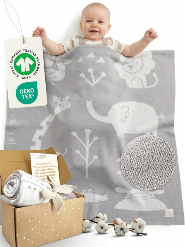 Glückliches Baby liegt unter grau-weißer gestrickter Babydecke aus Baumwolle mit OEKO-TEX und GOTS Anhänger – hochwertige Bio Baumwolle Babydecke, zusammengerollt auch in Geschenkbox präsentiert, ideal als Meilenstein Decke für Babys.