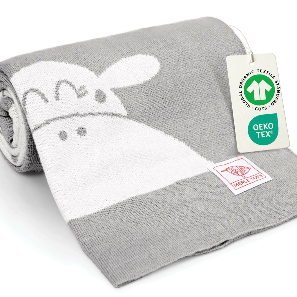 Grau-weiße gestrickte Babydecke aus Baumwolle, zusammengerollt mit OEKO-TEX und GOTS Anhänger – hochwertige Bio Baumwolle Babydecke, ideal als Meilenstein Decke für Babys.