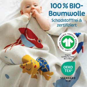 Ein Baby kuschelt sich in die Merle Toys Baumwoll Babydecke mit fröhlichen Meeresmotiven. Die gestrickte Babydecke besteht aus 100 % Bio-Baumwolle, ist schadstofffrei und zertifiziert. Durch ihre hochwertige Qualität ist sie ein schönes Geschenk zur Taufe Mädchen und als leichte gestrickte Babydecke Sommer angenehm weich auf empfindlicher Babyhaut.