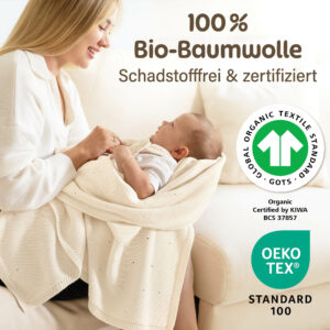 Eine lächelnde Mutter hält ihr Baby auf dem Sofa in einer Weisse Babydecke von Merle Toys, mit Hinweis auf 100 % Bio-Baumwolle, GOTS und OEKO-TEX Standard 100; die Babydecke-Strick wirkt weich, schadstofffrei zertifiziert und als kuschelige Baumwoll Babydecke ideal für den Sommer.