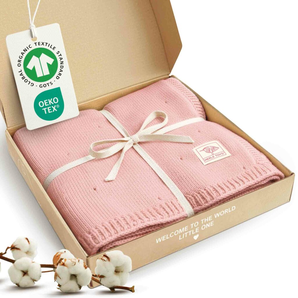 In der Geschenkbox liegt die Babydecke Strick von Merle Toys in Rosa, gebunden mit Schleife und ergänzt durch GOTS- sowie OEKO-TEX-Hinweise – eine hochwertige Babydecke Rosa aus Baumwolle, die sich als Sommerdecke für Babys und Geschenk zur Geburt Mädchen anbietet.