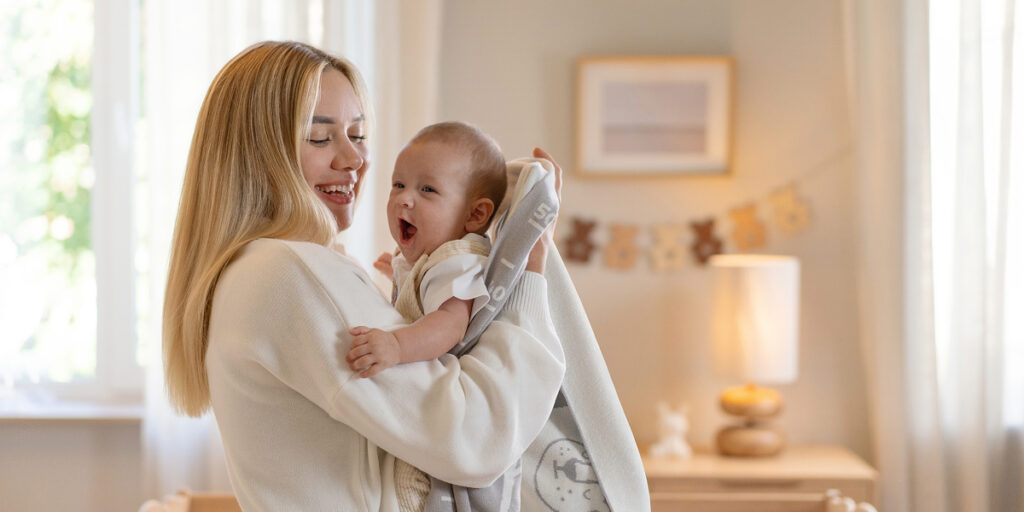 Merle Toys – Mutter hält ihr Baby im Arm, eingehüllt in eine perlweiße, gestrickte Kuscheldecke aus Bio-Baumwolle.