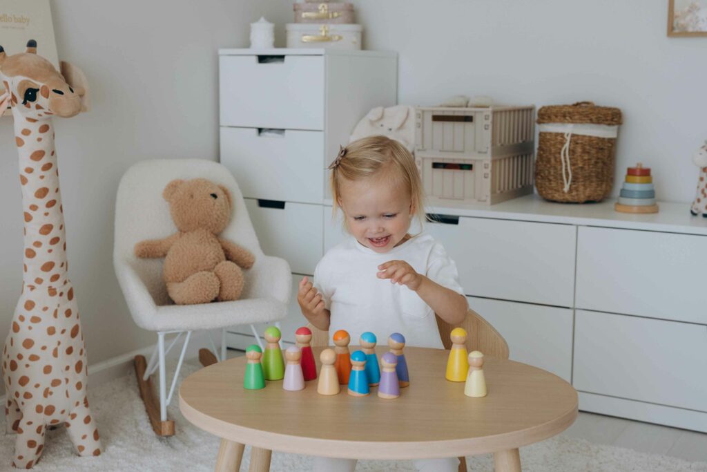 Kleines Mädchen spielt am Tisch mit farbigen Holz-Regenbogenfiguren – ein pädagogisch wertvolles Lernspiel und ideales Geschenk für 2-Jährige. Perfekt als sinnvolles Geschenk für 2-Jährige für Jungs & Mädchen und beliebt als Weihnachtsgeschenk für 2-Jährige.