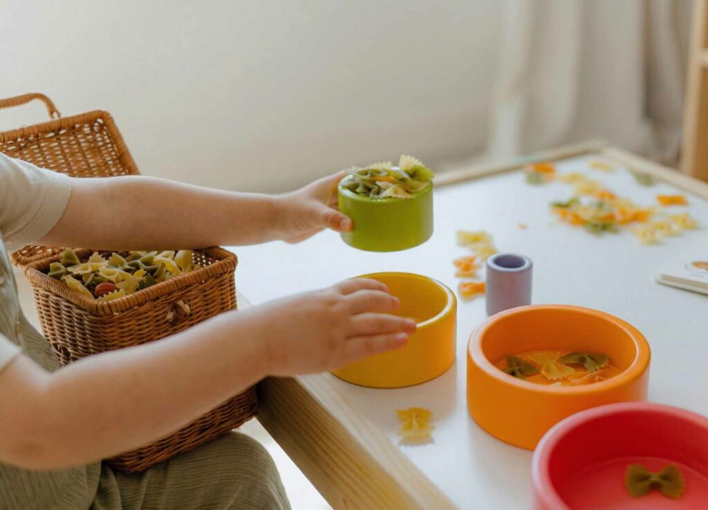 Kind sortiert kleine Nudeln in bunte Schalen und trainiert spielerisch den Pinzettengriff – ideal, um die Feinmotorik zu fördern und zu zeigen, ab wann Babys gezielt greifen.