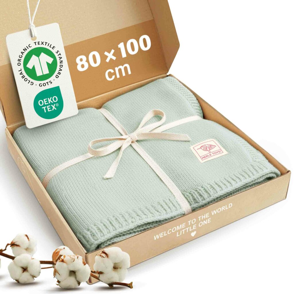 Eine mintgrüne Babydecke-Strick von Merle Toys liegt gefaltet mit Schleife in einer Geschenkbox, daneben GOTS- und OEKO-TEX-Siegel, Baumwollzweige und die Größe 80 x 100 cm; die Babydecke Baumwolle Bio eignet sich ideal als Babydecke für Sommer.