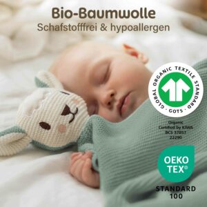 Ein schlafendes Baby liegt neben der grünen Merle Toys_Knuddeldecke mit angenähtem Schafsköpfchen. Die Baby Kuscheldecke aus Bio Baumwolle wird mit GOTS und OEKO TEX Kennzeichnung gezeigt und ist als gestrickte Babydecke Grün, Babydecke Baumwolle und Kuscheldecke für Babys gedacht.