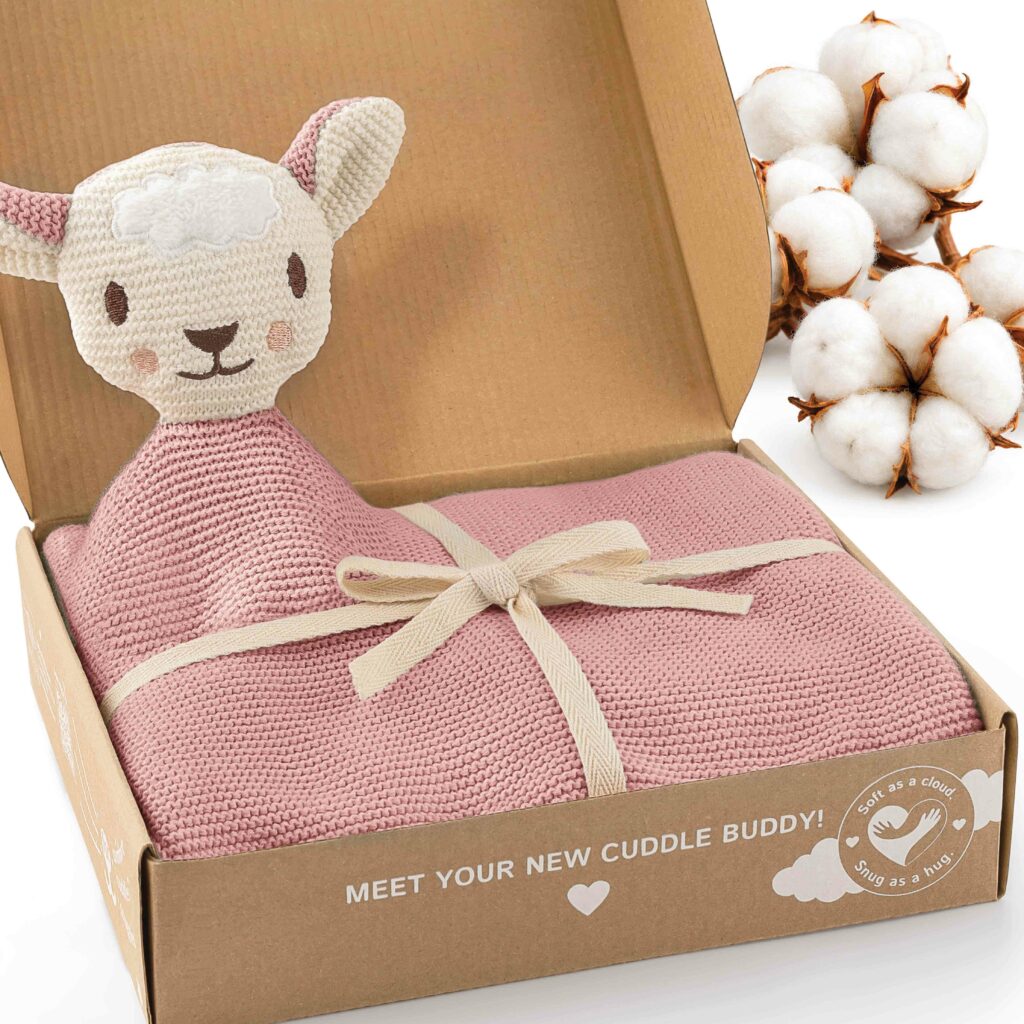 Geöffnete Geschenkbox mit rosa gestrickter Babydecke und angenähtem Schafsköpfchen neben Baumwollblüten. Die Merle Toys_Knuddeldecke ist eine weiche Baby Kuscheldecke Mädchen und eignet sich als Babydecke Strick, Baumwoll Babydecke Winter und Geschenk für Neugeborene.