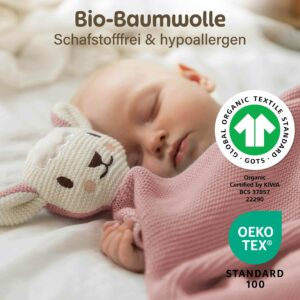 Ein schlafendes Baby liegt neben der rosa Kuscheldecke Baby mit süßem Schafsköpfchen. Im Bild sind außerdem die GOTS und OEKO TEX Zertifizierungen der Merle Toys_Knuddeldecke zu sehen. Die Baby Kuscheldecke Mädchen aus Baumwolle eignet sich auch als Baumwoll Babydecke Winter und Geschenk für Neugeborene.