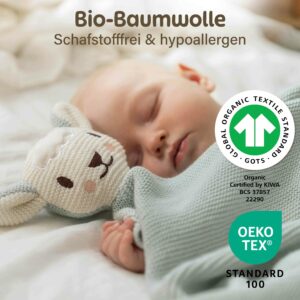 Ein schlafendes Baby liegt neben der mintfarbenen Merle Toys_Knuddeldecke mit Schafsköpfchen. Die Kuscheldecke für Babys aus Bio Baumwolle wird mit GOTS und OEKO TEX Kennzeichnung gezeigt und eignet sich als Baby Decke für Neugeborene, Kinderwagen Decke, Baumwolldecke und Babydecke Winter sowie als Geschenk zur Geburt Mädchen.