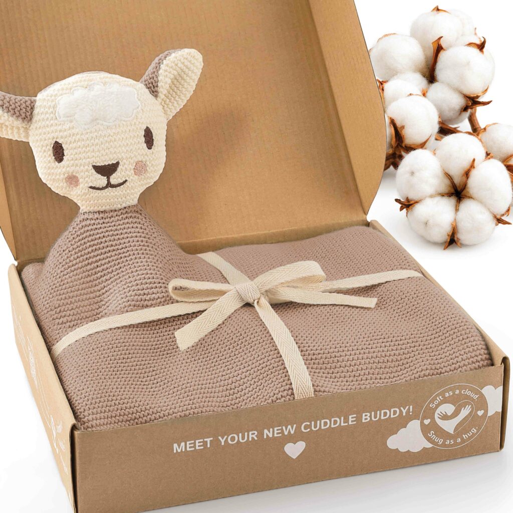 Geöffnete Geschenkbox mit der beige braunen Merle Toys_Knuddeldecke, angenähtem Schafsköpfchen und dekorativen Baumwollblüten. Die weiche Kuscheldecke für Babys ist eine gestrickte Babydecke Winter und Sommer und eignet sich als Strick Babydecke für den Kinderwagen sowie für Baby Kuscheldecken Mädchen und Jungen.