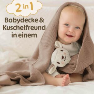 Ein lächelndes Baby sitzt eingekuschelt in die beige braune Merle Toys_Knuddeldecke und hält das integrierte Schafsköpfchen nah bei sich. Die 2 in 1 Kuscheldecke für Babys verbindet Kuschelfreund und Babydecke Winter und Sommer und eignet sich auch als Strick Babydecke für den Kinderwagen sowie für Baby Kuscheldecken Mädchen und Jungen.